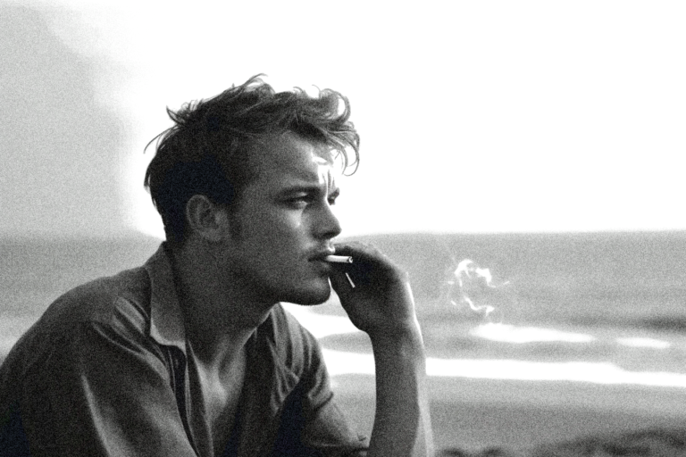 a young male artist is laying on a beach smoking a cigarette